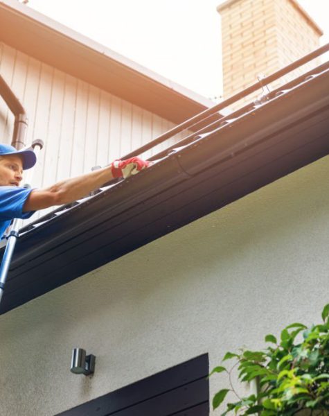 man on ladder cleaning house gutter from leaves and dirt