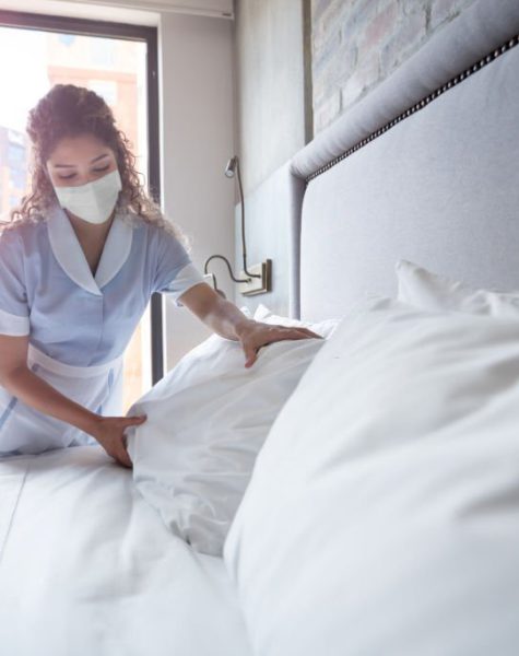 Latin American maid working at a hotel and doing the bed wearing a facemask - COVID-19 pandemic lifestyle concepts