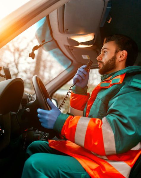 An adult handsome male paramedic is talking on a portable radio while sitting in an ambulance outside a clinic.